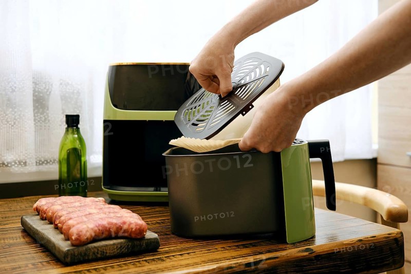 Person places parchment paper under crisper tray in food basket of air fryer in kitchen. Fresh sausage sits on wooden cutting board. Olive oil bottle present on table next to air fryer