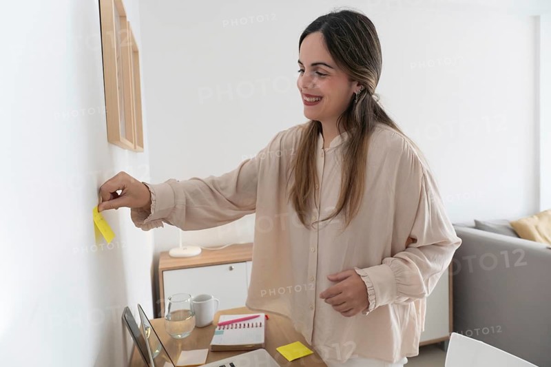 Smiling content creator placing yellow sticky note on wall next to framed print, desk with laptop and notebook visible in bright apartment