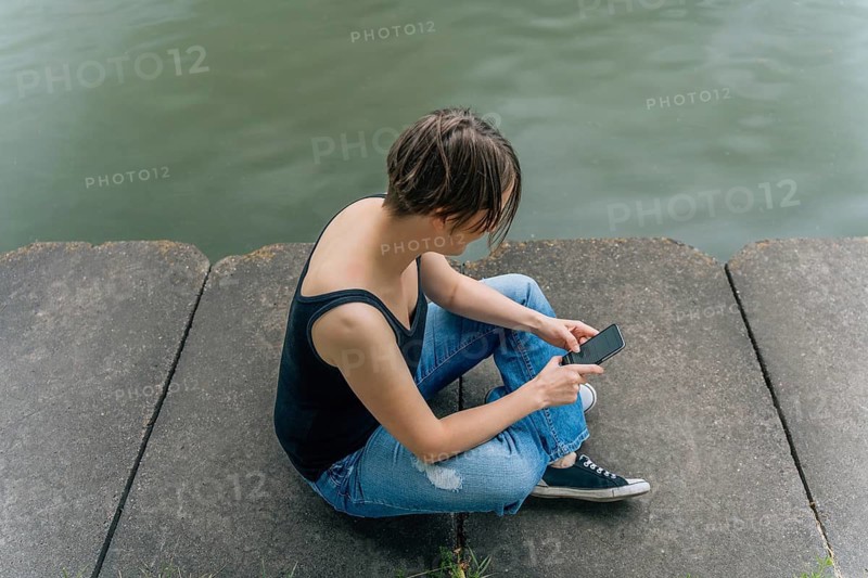 Woman Using Smartphone Sitting by the Water's Edge, A person sits on a concrete surface by the water, using a phone, appearing relaxed and casual