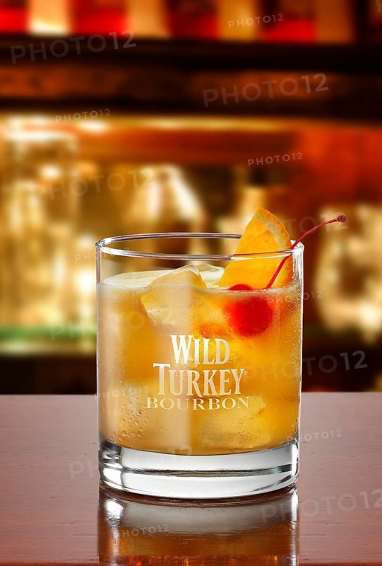 Whisky on ice cube cubes in a tumbler on a bar counter in front of a bar background, cocktail cherry and half an orange slice as a garnish, studio recording