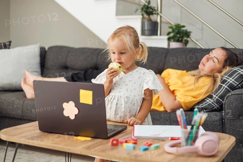 Young blond European girl eating an apple near a laptop on a table. Relaxed European woman with dark hair lying on a sofa in the background