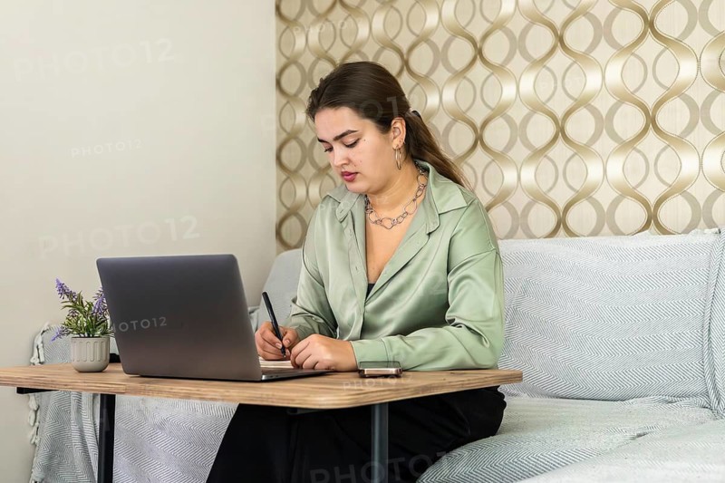 A woman sits at a table with a laptop in front of her. She writes notes while focused on her task. The room has a light design with patterned walls. It's daytime and the atmosphere is active