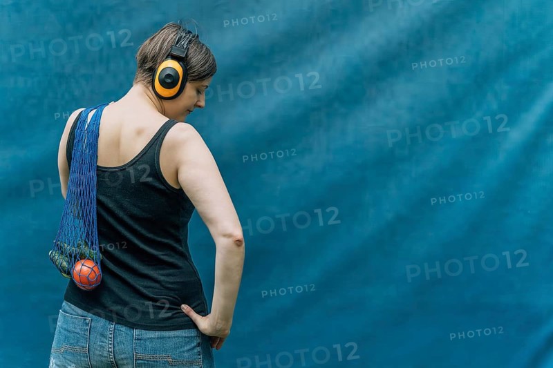Woman with yellow headphones carrying mesh grocery bag, back view on blue, A woman with headphones, wearing a tank top and jeans, carries a net bag with a ball against a blue background