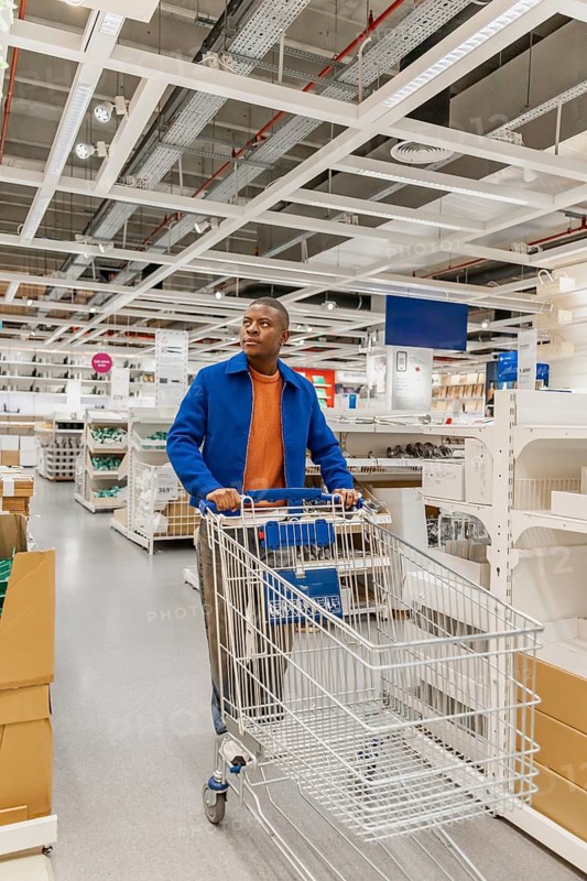 A young Black man pushes a shopping cart in a modern retail store. The interior features bright lighting and organized shelves filled with products