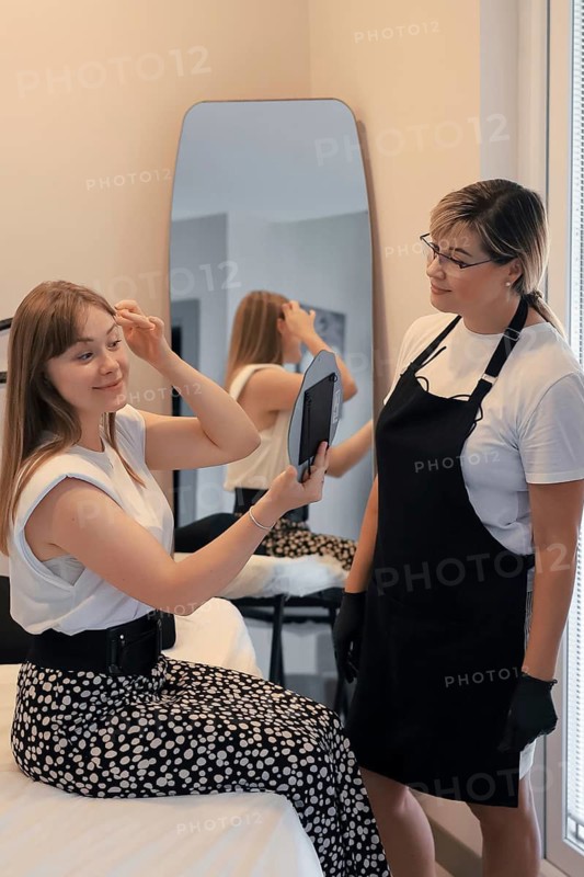A woman checks her makeup in the mirror while the stylist observes