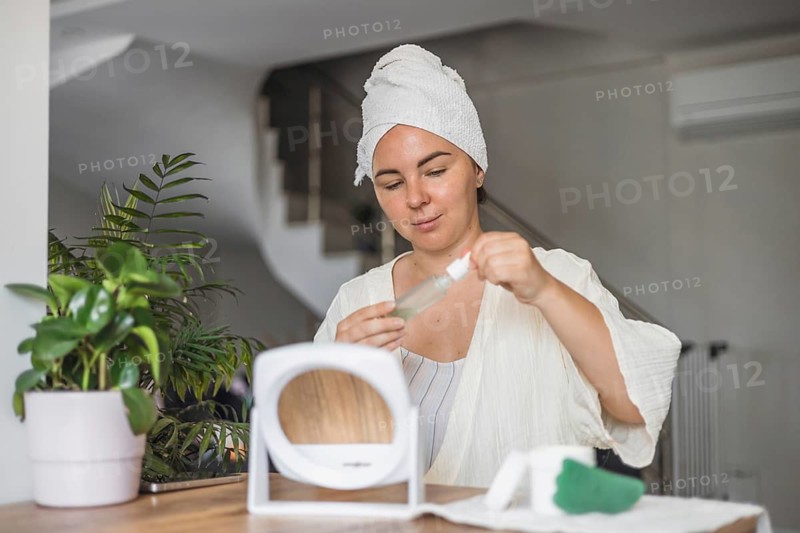 A young Caucasian woman with brown hair in a towel prepares for a gua sha face massage at home. She holds a serum bottle near a mirror and plants