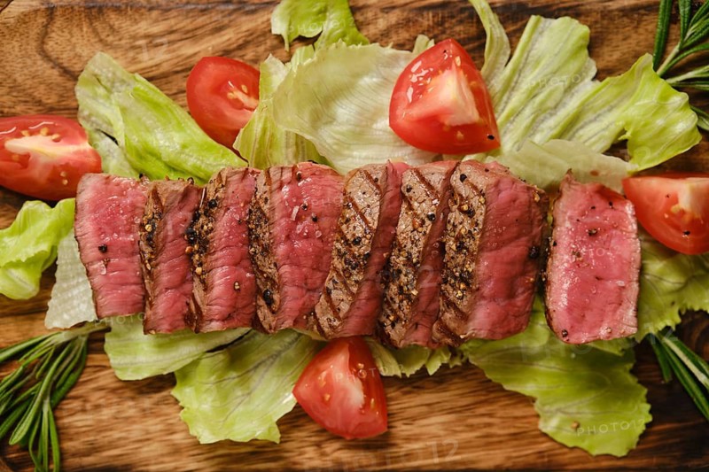 Beef steak is arranged neatly on wooden plate. Fresh lettuce and sliced tomatoes surround steak. This meal is prepared in a kitchen setting, ready to be served at dinner time