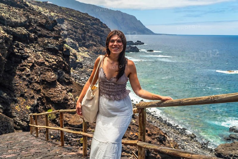 Young woman smiling while standing on a path with a wooden handrail, overlooking the deep blue ocean and rugged volcanic coastline of el hierro in the canary islands