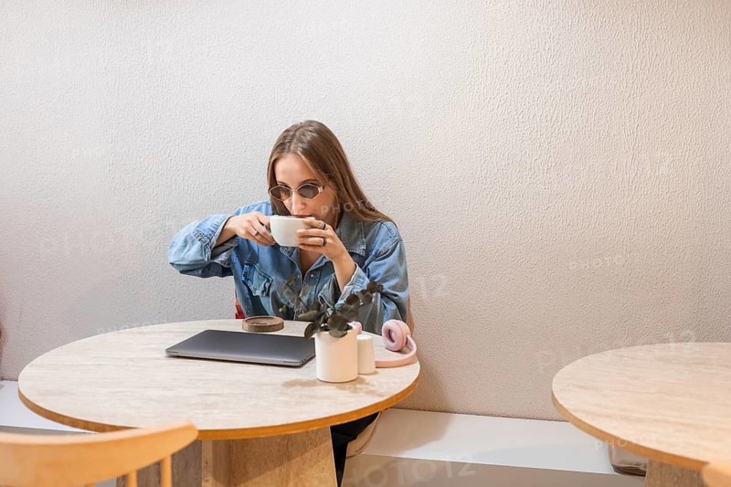 A young Caucasian woman with long brown hair sits at a wooden table, sipping coffee from a white cup. A laptop and a small plant are on the table
