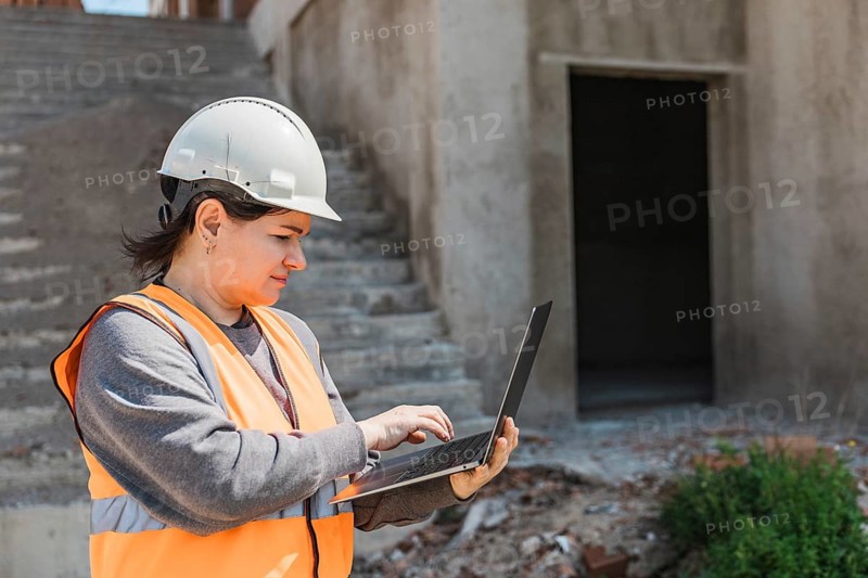 Woman in a hard hat and orange vest uses a laptop on a construction site. Stairs and an unfinished doorway are visible in the background