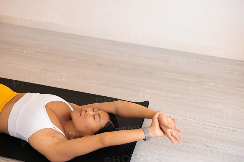 A woman is lying on a yoga mat with her arms relaxed above her head in a serene posture