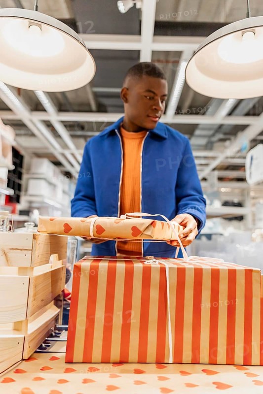 A young Black man with short hair stands at a table in a store, wrapping gifts in colorful paper. The setting is bright and modern, with shelves in the background