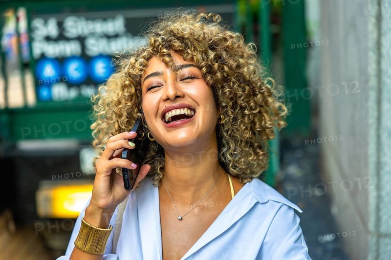 Happy young woman with curly hair laughing while having a phone call, standing in front of a manhattan subway entrance in new york city, enjoying urban lifestyle
