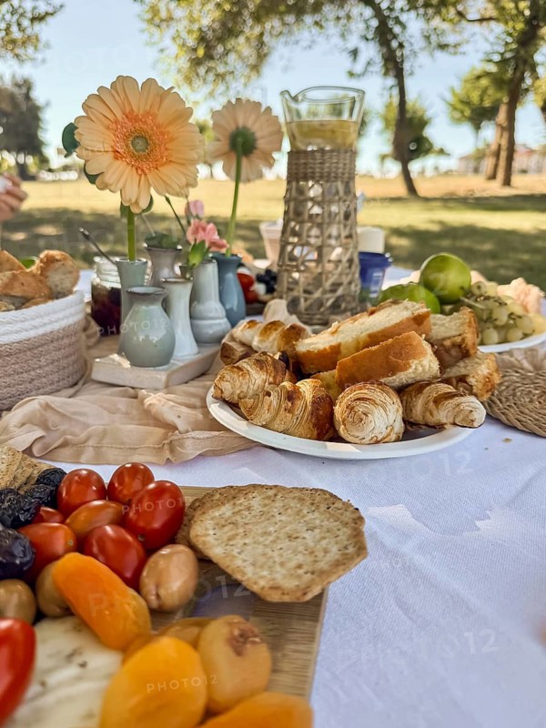 A picnic spread featuring fresh fruits, bread, and pastries on a white tablecloth. Flowers and drinks are arranged in a sunny outdoor setting