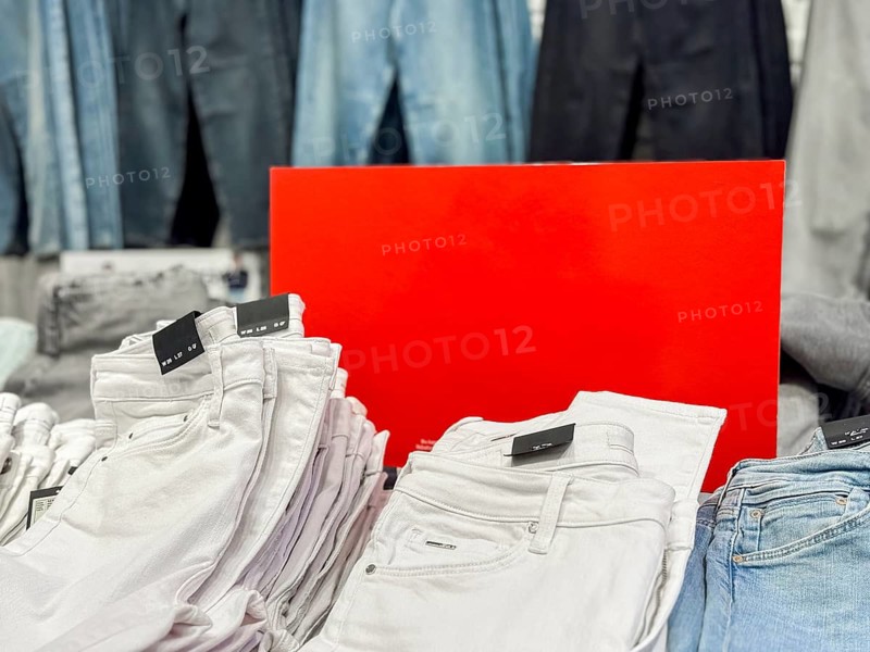 A display of folded white jeans in a retail store. Behind them, various shades of blue denim pants hang on racks. A bright red sign is positioned in the background