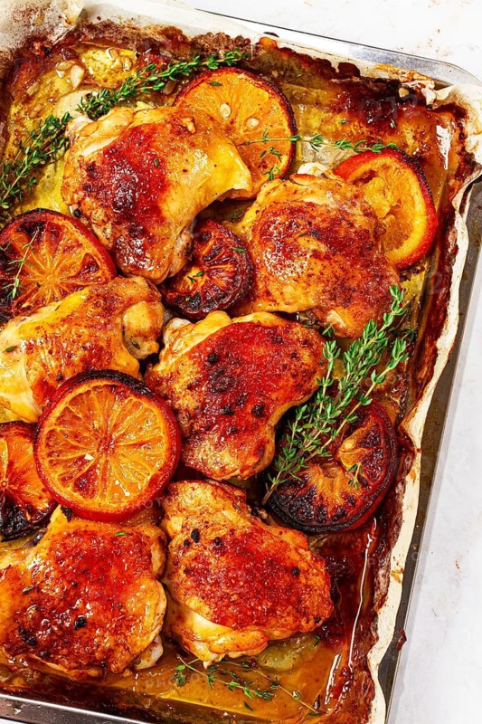 A tray holds cooked chicken thighs topped with orange slices and fresh herbs. The chicken has a rich brown color and is surrounded by juices. This scene shows a kitchen ready for a meal