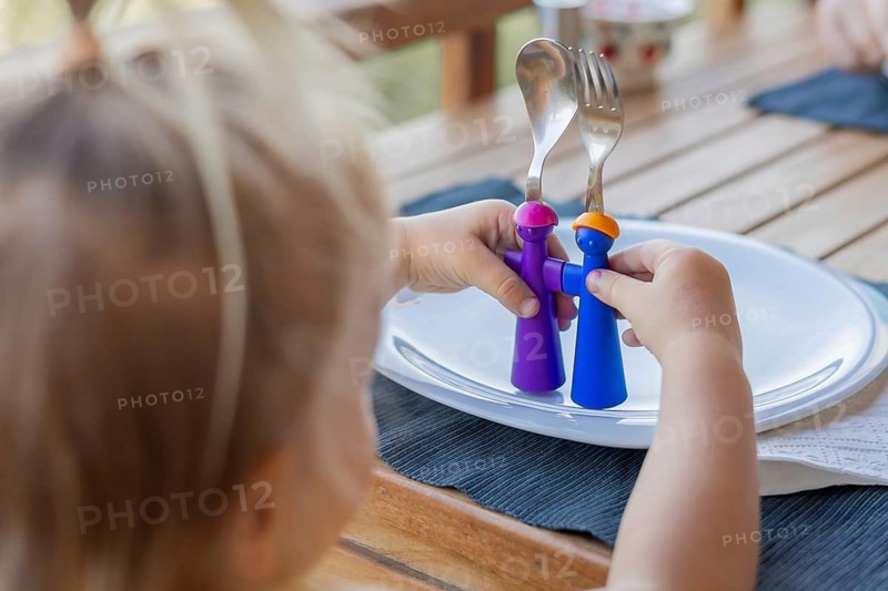 Young child interacts with colorful utensils, engaging in imaginative play at a wooden table during a family gathering