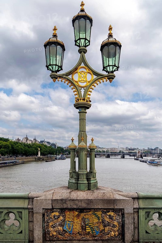 A three headed cast iron lamp post stands on Westminster Bridge above a heraldic panel, part of the bridges original Victorian decoration. Opened in 1862 with Gothic ornamentation and royal symbolism, the bridge forms a ceremonial approach to the Houses of Parliament, London, England, United Kingdom