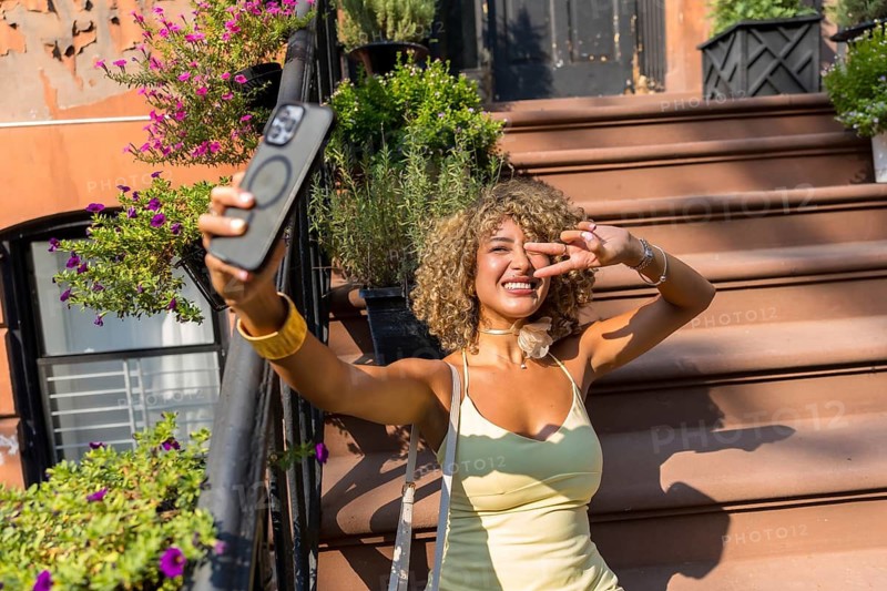Happy young woman with curly hair taking a cheerful selfie on the steps of a brownstone building in manhattan, capturing a vibrant urban summer lifestyle