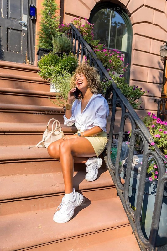 Young woman with curly hair sitting on a brownstone stoop, laughing while talking on her cell phone, enjoying city lifestyle on a sunny day in new york