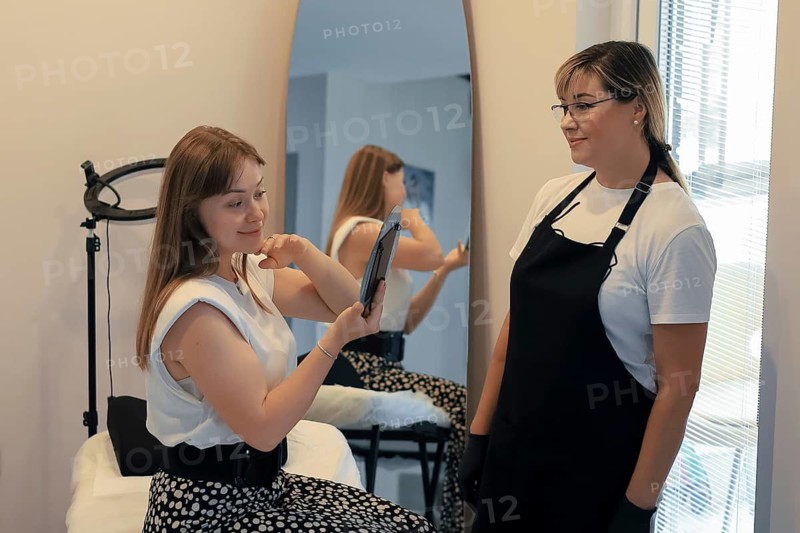 A woman examines her makeup in a handheld mirror in the presence of a stylist