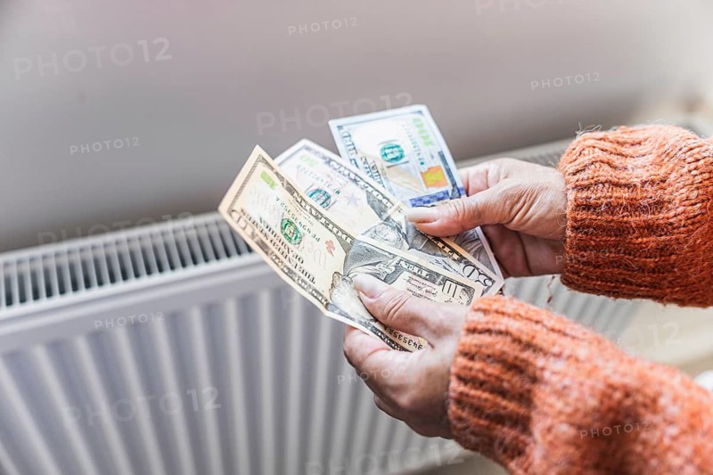 Winter home heating expenses and rising utility costs. Woman counting US dollar cash near radiator as concept of inflation, household energy spending and cost of living pressure.