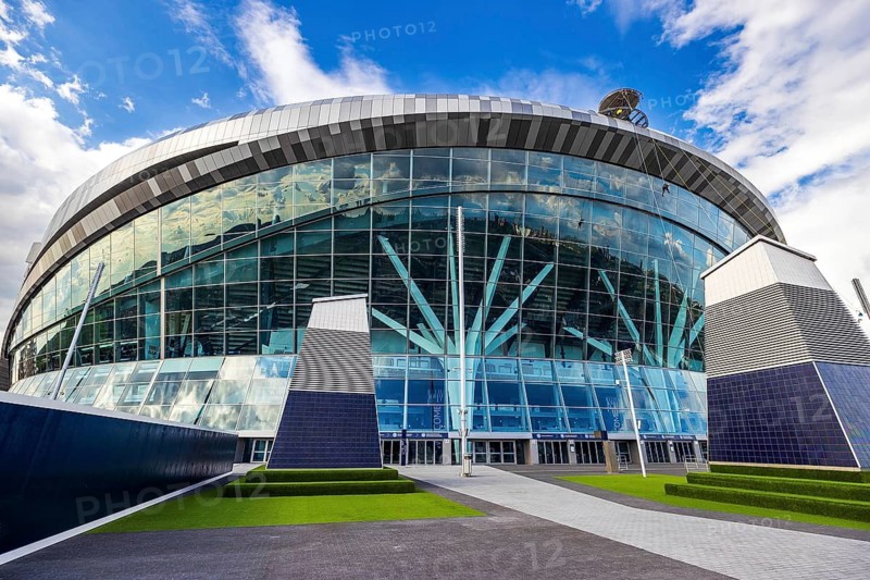 Landscaped forecourt leads to the glazed entrance of Tottenham Hotspur Stadium on a clear day, London, England, United Kingdom