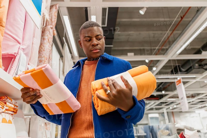 A young Black man examines colorful towels in a retail store. He holds an orange towel in one hand and a pink striped towel in the other, contemplating his purchase