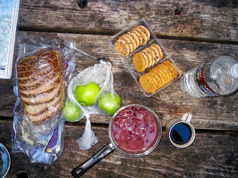 View from above of a rustic camping table with bread, apples, cookies, kidney beans in a cooking pot, bottle of water, cup of coffee, emergency food, picnic, meager breakfast outdoors, USA