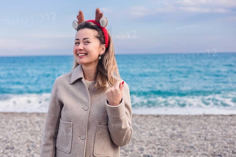 A young Caucasian woman with brown hair wears reindeer antlers at the beach during winter. The ocean is visible in the background, creating a festive atmosphere