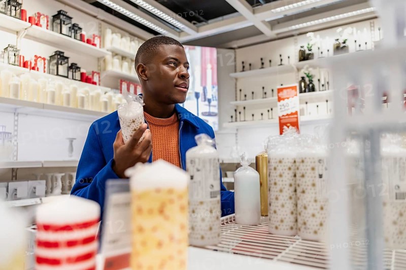 A person explores a store that sells candles, holding one candle while looking at many other candles displayed on shelves. The setting is bright and organized with various products