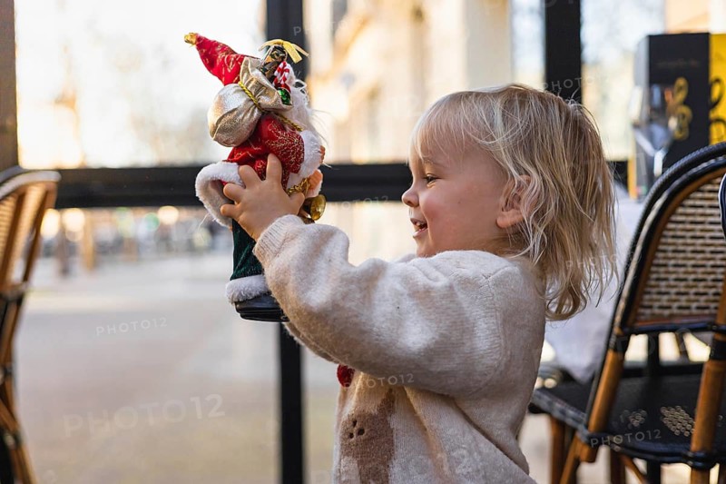 A young Caucasian girl with blonde hair smiles while holding a Santa Claus figurine. The scene is festive, capturing the joy of Christmas celebrations