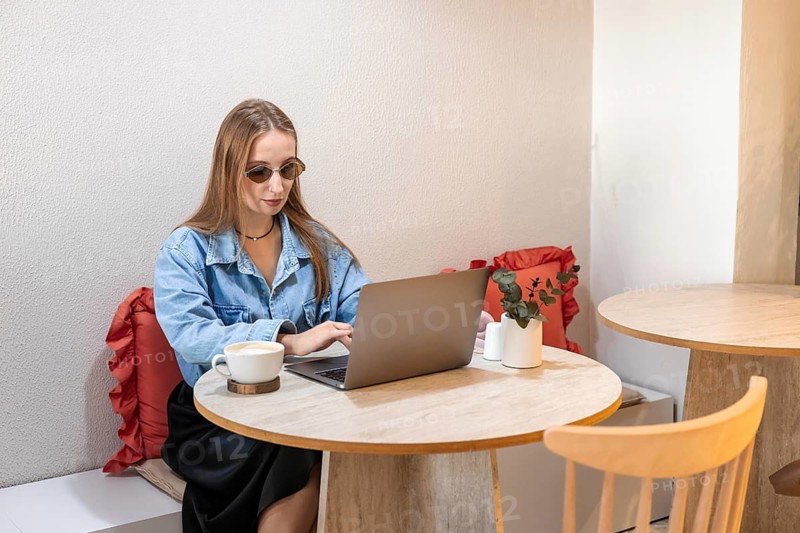 Young Caucasian woman with long brown hair wearing sunglasses, working on a laptop at a table with a plant and a cup