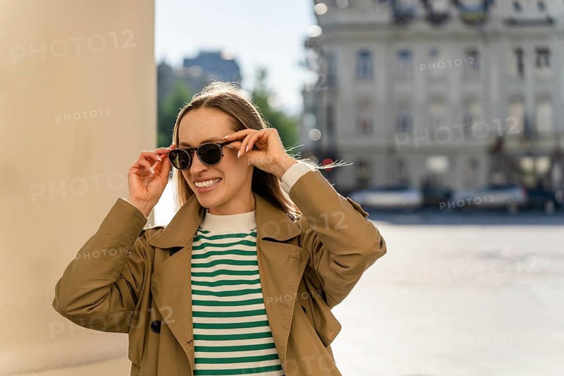 Smiling woman puts on sunglasses outside in the city, A woman in a trench coat and sunglasses smiles in a sunny urban setting, Vilnius, Lithuania