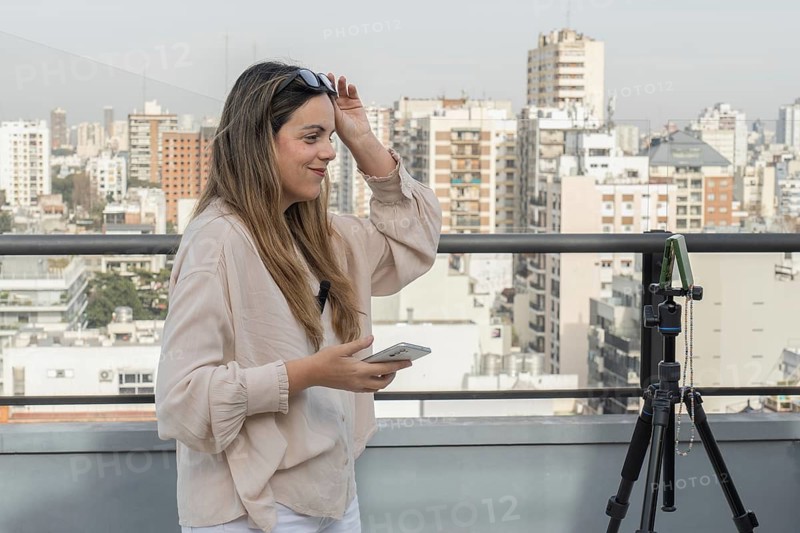 Smiling woman lifting sunglasses off her head, holding smartphone near tripod, city skyline spreading behind rooftop railing
