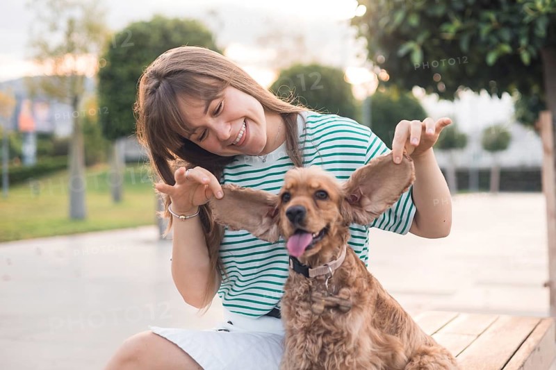 A woman playfully laughs with her dog, both sitting on a park bench
