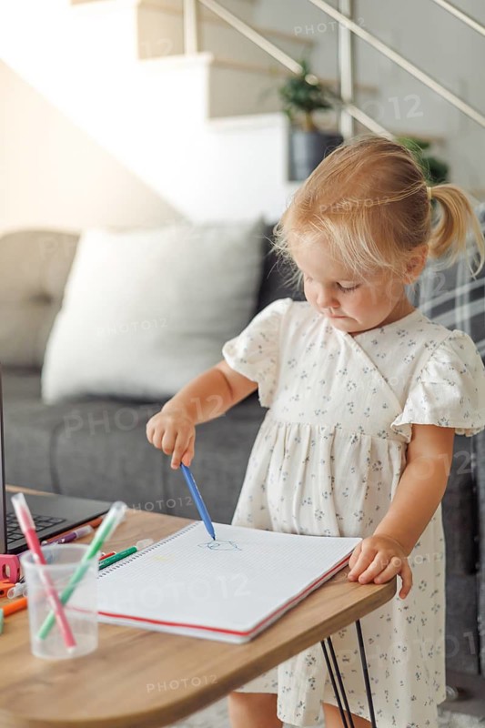 Young blond European girl drawing with colored pencils at a table in a living room with a laptop and toys