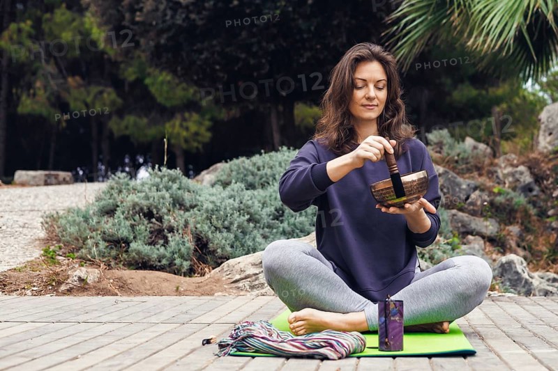 A young Caucasian woman with brown hair sits cross-legged on a yoga mat outdoors. She is using a singing bowl, surrounded by greenery and wooden decking