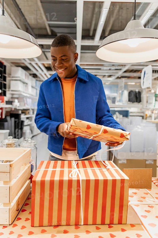 A young Black man smiles while holding a gift box in a modern shopping environment. Colorful packaging and organized displays are visible around him