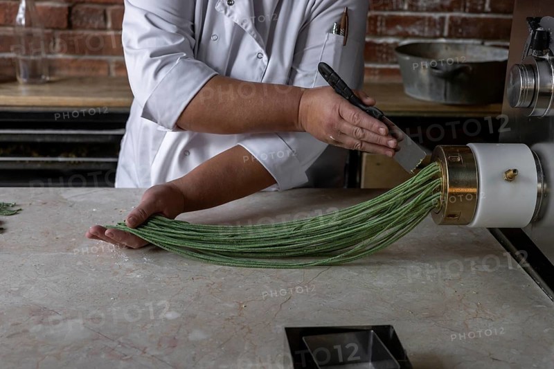A chef skillfully extrudes vibrant green noodles from a pasta machine in a modern kitchen. The bustling atmosphere reflects the lively lunch hour at the restaurant