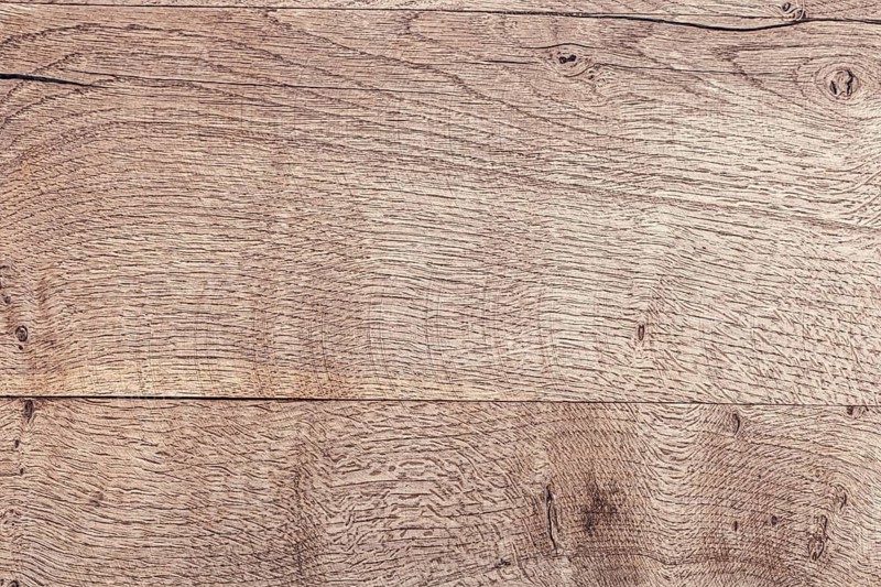 Close-up of a textured wooden surface with natural grain patterns. The wood is light brown with subtle variations in color and texture