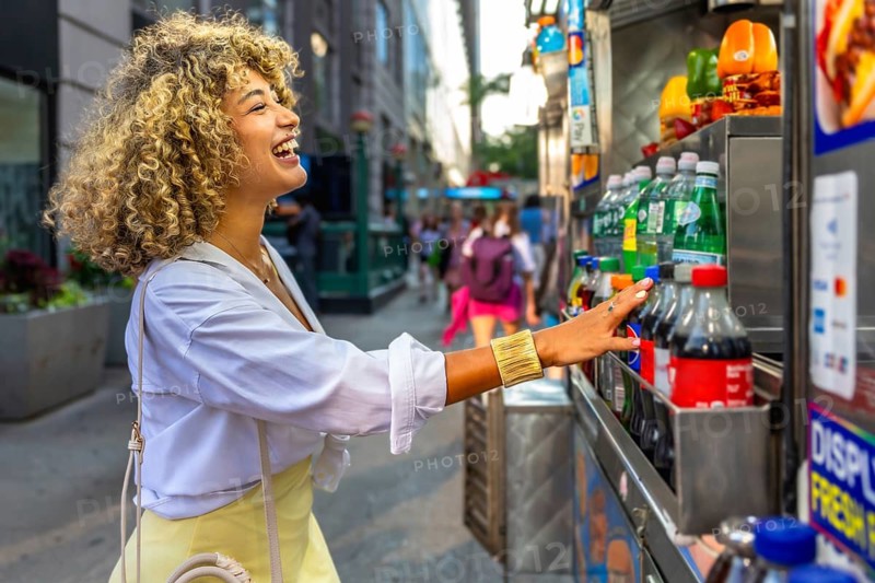 Woman with blonde curly hair enjoying street food culture in new york city, reaching for a beverage from a vendor cart on a busy manhattan street, embracing urban lifestyle and happiness