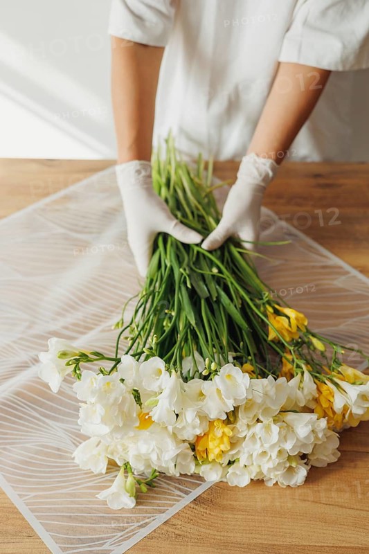 A person in gloves carefully arranges a bunch of fresh flowers, combining vibrant yellow and white blooms. The setting is a bright, airy workspace, enhancing the natural beauty of the flowers