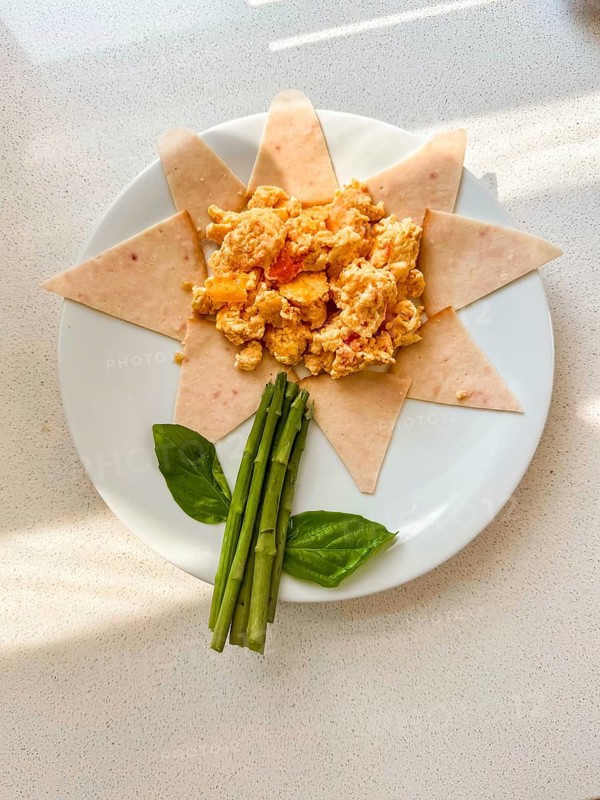 A creative children breakfast plate arranged like a flower. Scrambled eggs in the center, surrounded by tortilla slices, with asparagus and basil leaves as decoration