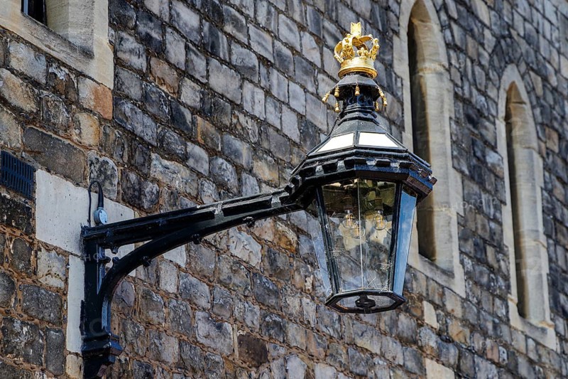 A black iron lantern topped with a gilded crown projects from a stone wall within Windsor Castle, one of many decorative lamps throughout the precinct that combine practical lighting with visible royal insignia, Windsor, England, United Kingdom