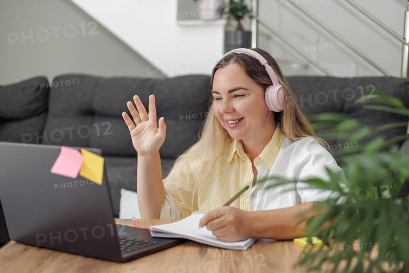 Young blond European woman with headphones waving hand during video call at home with laptop and notebook on table