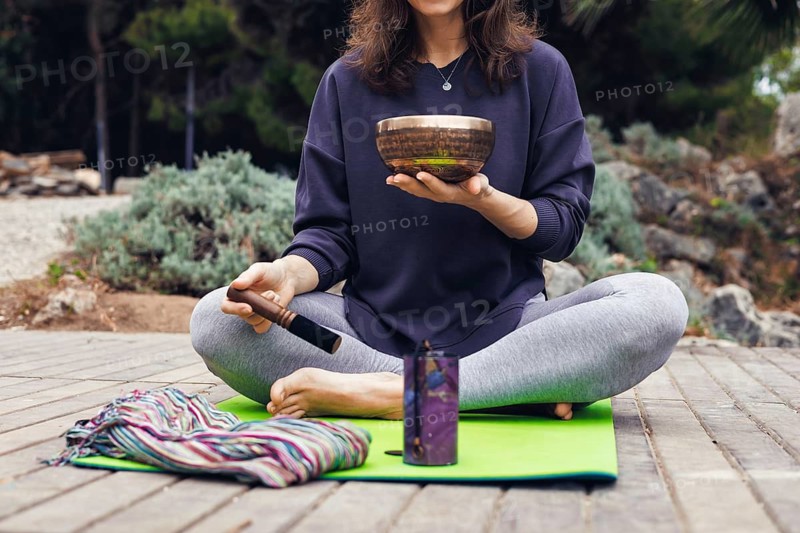 A young Caucasian woman with long brown hair sits cross-legged on a green yoga mat by a lake. She holds a singing bowl and appears to be meditating