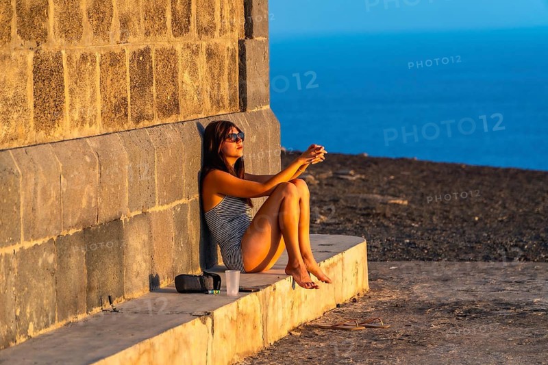 Woman resting against a stone wall, enjoying the warm light of the setting sun overlooking the ocean during a peaceful island vacation in the canary islands