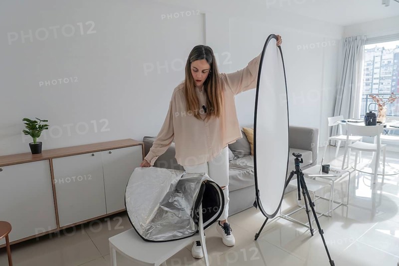 Woman standing and holding fully extended white photography reflector upright, city skyline visible through window, camera tripod nearby