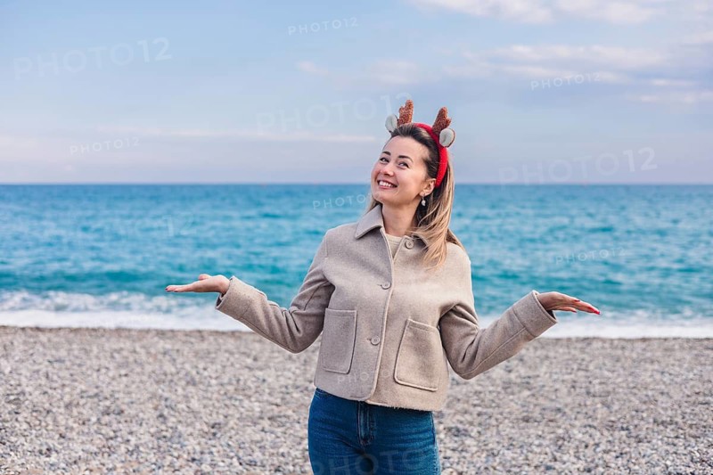 A young Caucasian woman with blonde hair wears reindeer antlers at a beach during winter. The ocean is visible in the background, creating a festive atmosphere